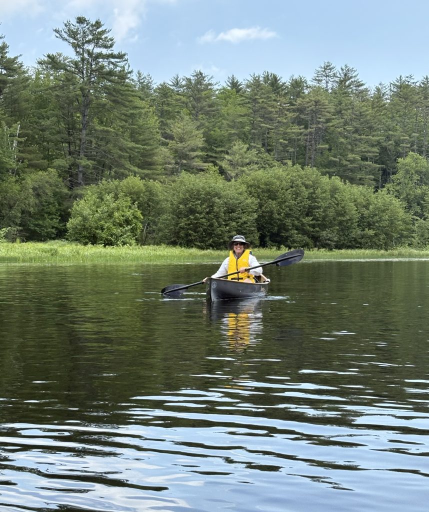 The photo shows Robin enjoying time outdoors in a kayak on  a river. We can see trees in the background and sparkling water around the boat. 